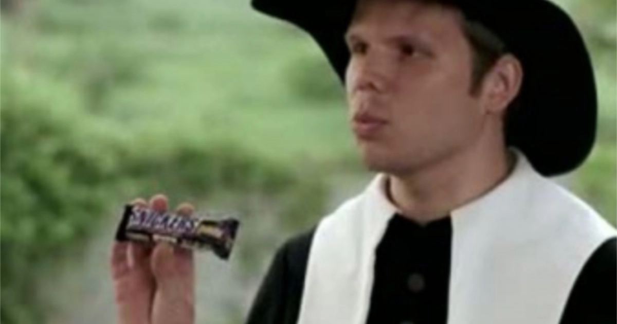 Commercial still of a man in a black hat holding a Snickers bar