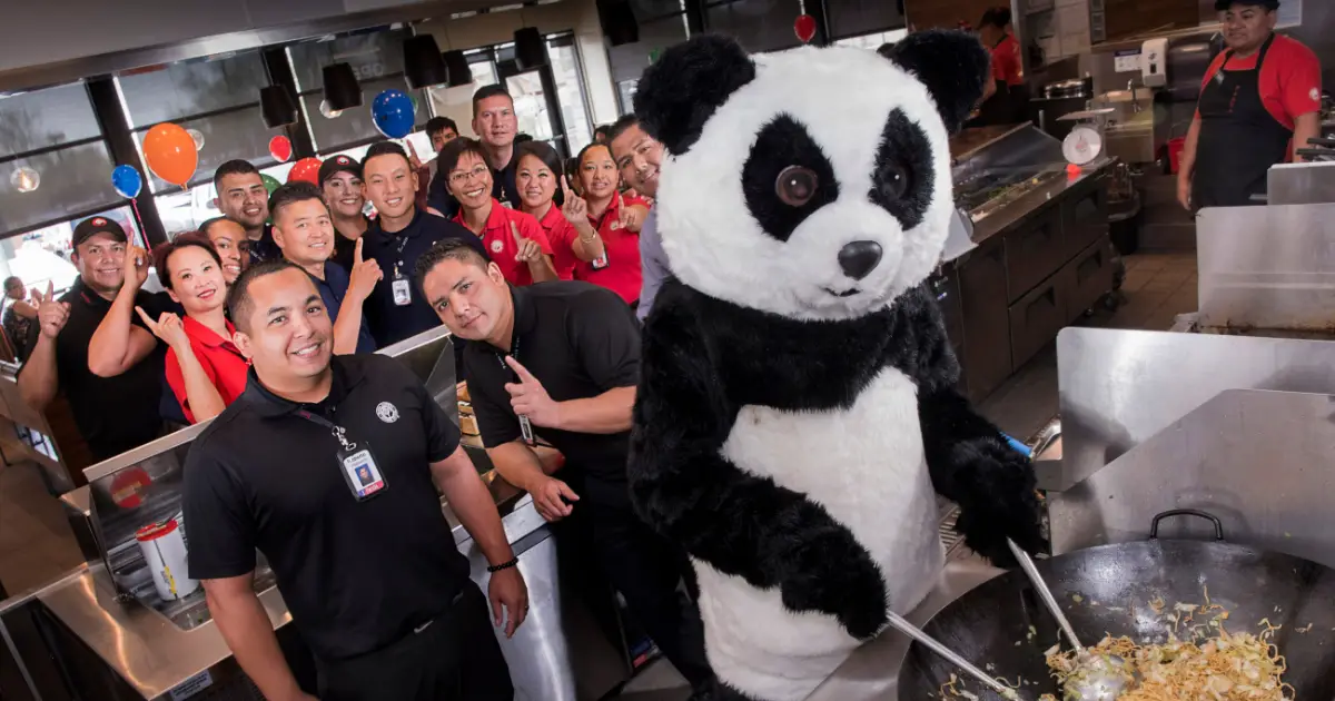 Chu with store crew at a Panda Express opening — Panda Express Panda Express employees posing with the Chu panda mascot costume in a restaurant kitchen