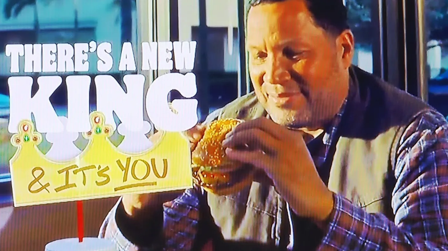Still from the 2026 Burger King commercial showing a customer eating a Whopper beside a paper crown and the words There's A New King and It's You