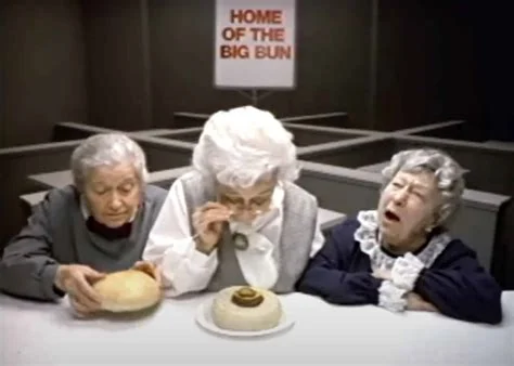Clara Peller and her co-stars size up the competition's bun in the 1984 "Fluffy Bun" spot — Wendy's Three elderly women examine a tiny burger patty on an oversized bun beneath a HOME OF THE BIG BUN sign in Wendy's 1984 Where's the Beef commercial